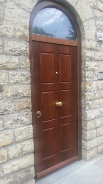 Porta d'ingresso ad arco in legno incastonata in un muro di pietra, con ferramenta in ottone e sopraluce in vetro satinato.