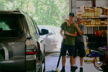 Two men stand in a garage. One has an arm around the other, holding a cleaning tool near a gray SUV.
