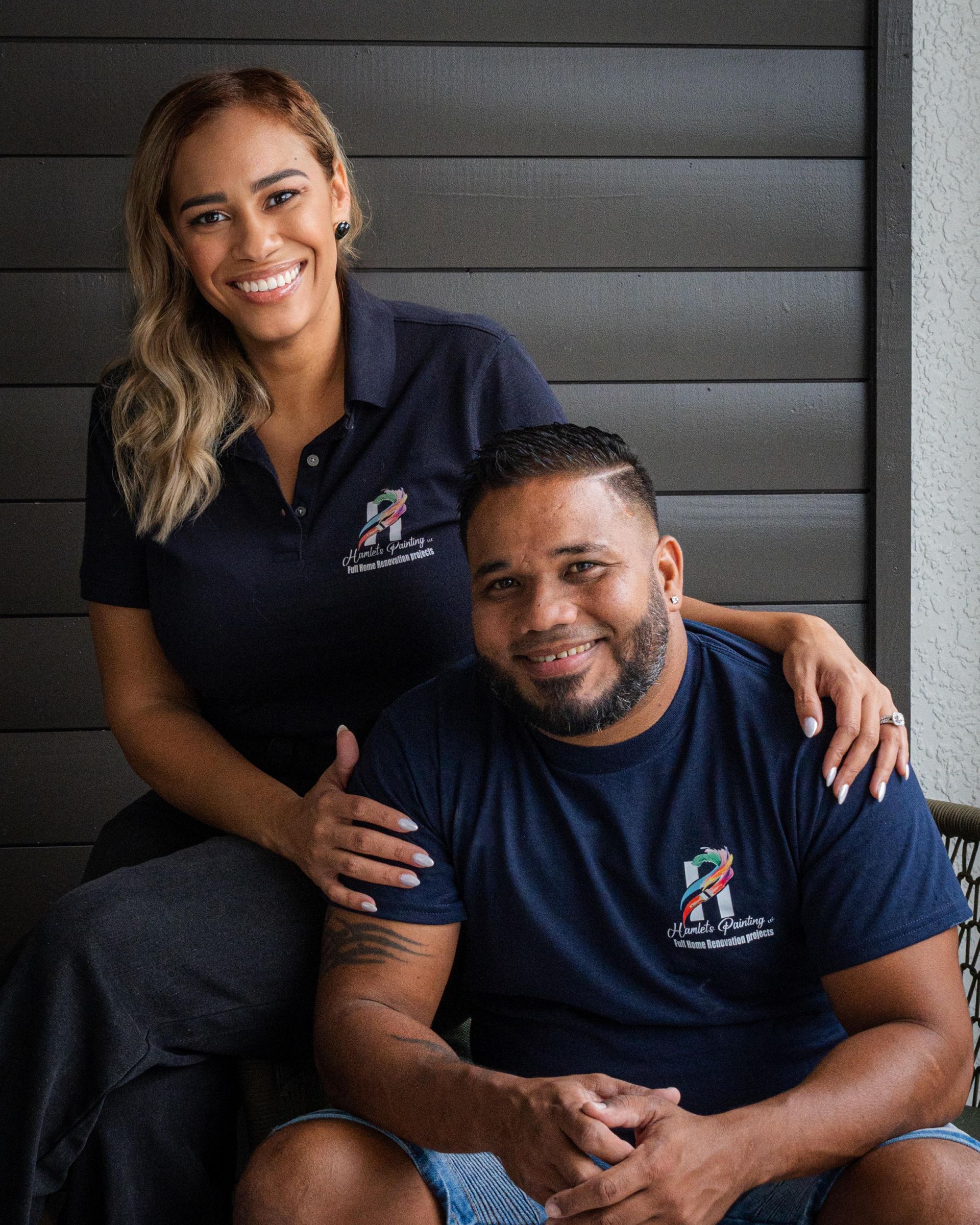Two people in matching navy shirts sit and smile against a dark paneled backdrop.
