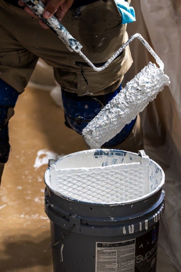 Painter rolling white paint onto a bucket lid with a gray paint bucket below.