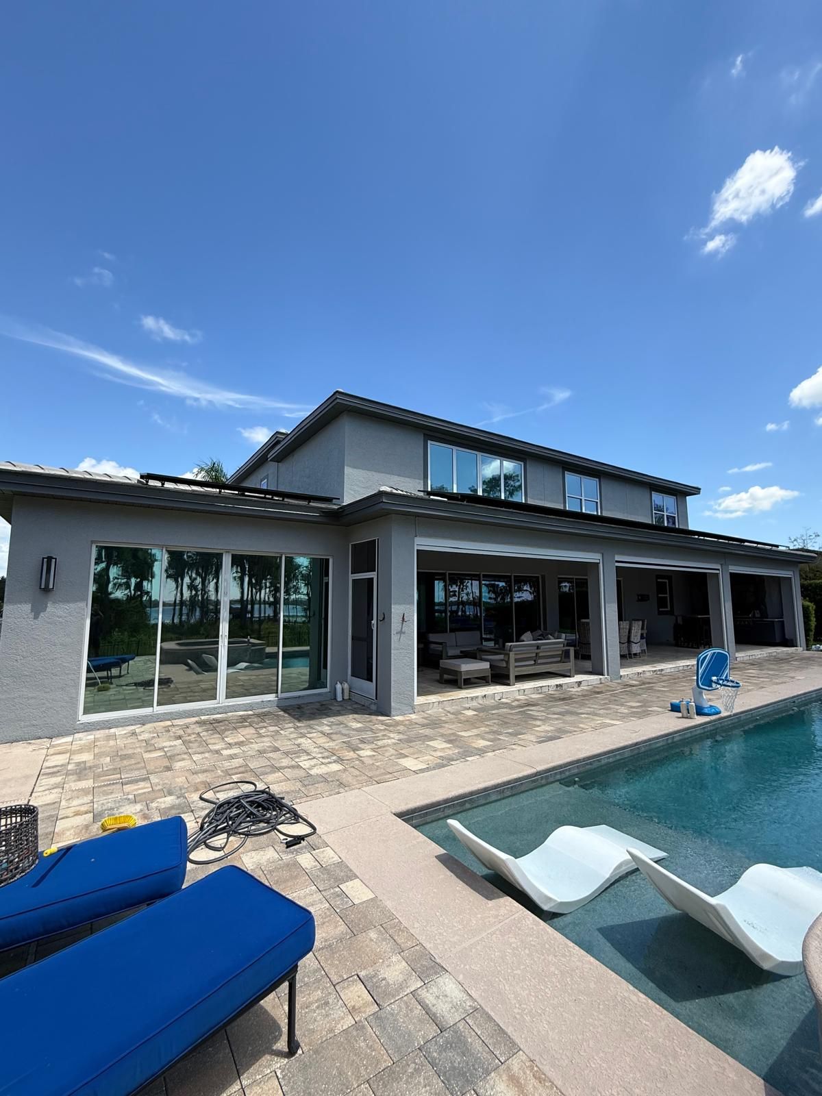 Modern house with covered patio and blue-cushioned lounge chairs beside a swimming pool under a clear sky