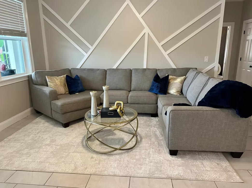 Gray sectional sofa in a bright living room with a geometric accent wall, rug, and glass coffee table.