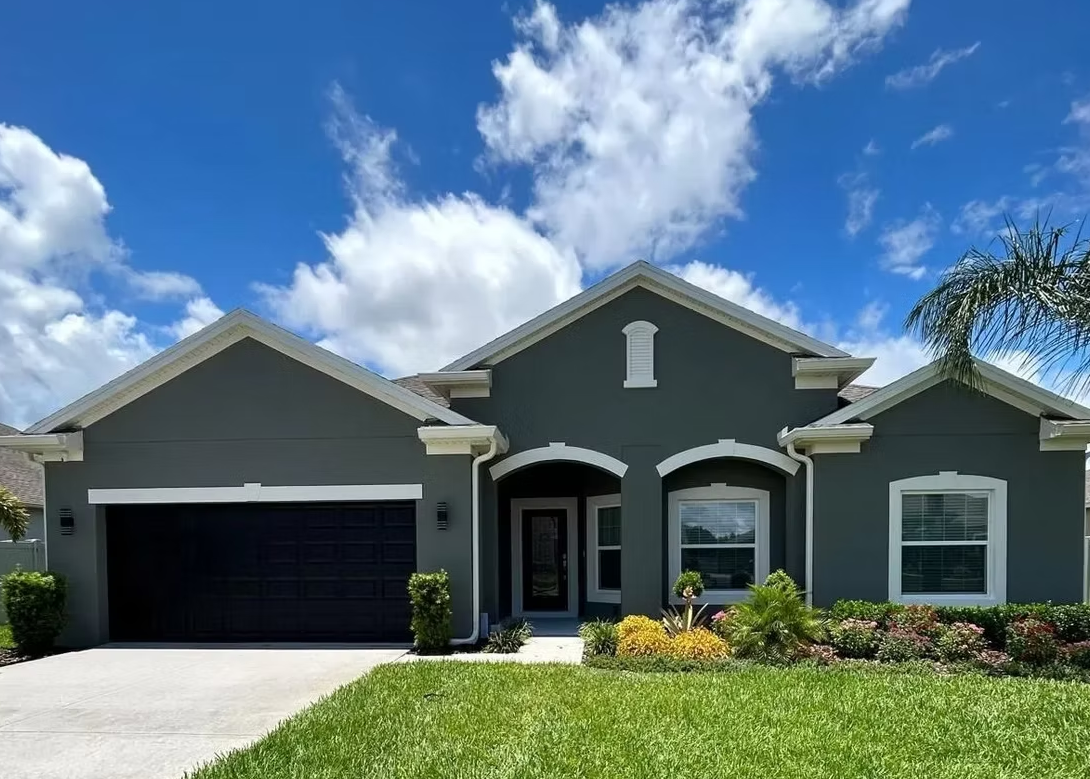 Gray suburban house with a black garage door, green lawn, and blue sky with clouds