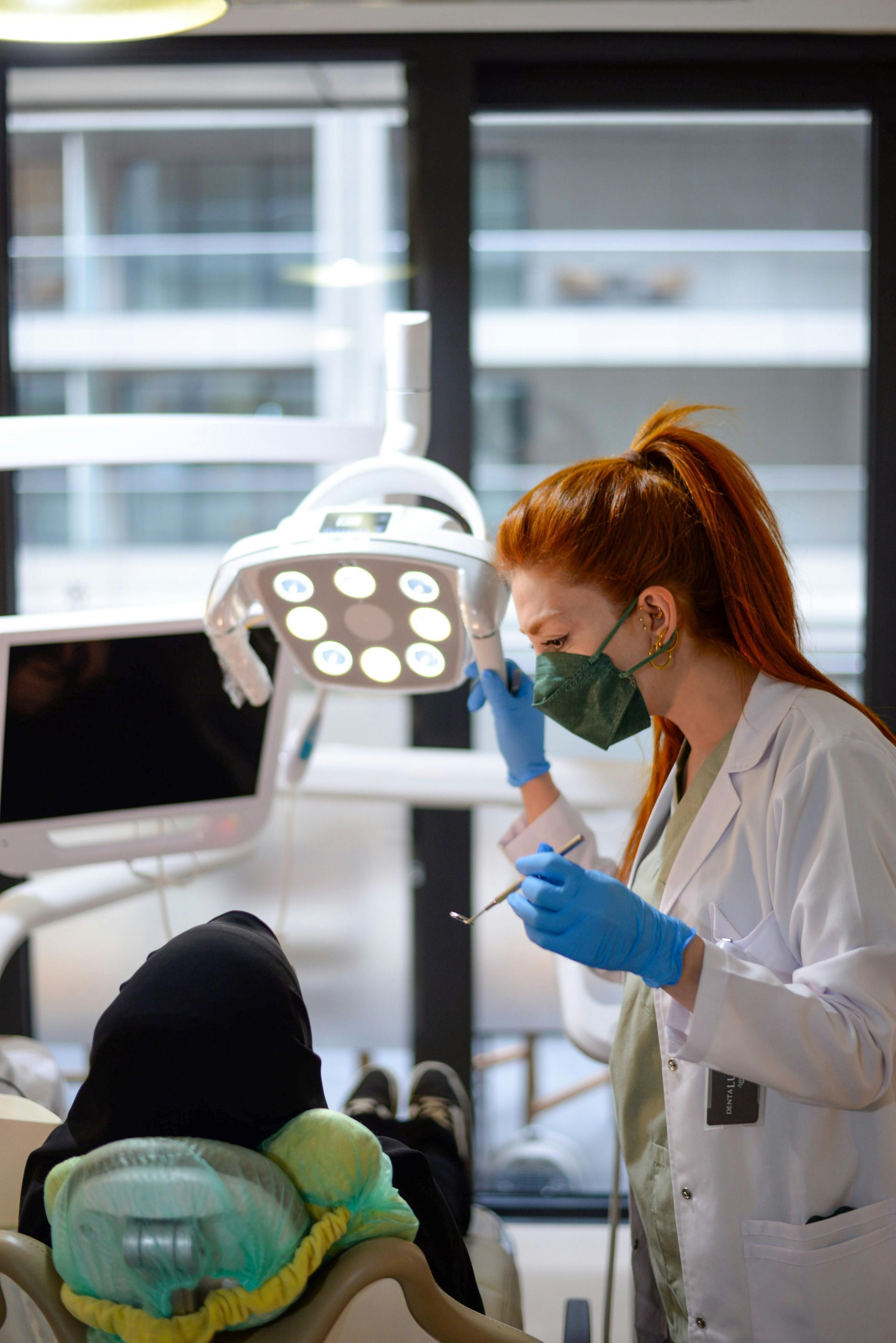 Dentist in white coat and mask examining a patient in a dental chair under bright exam lights