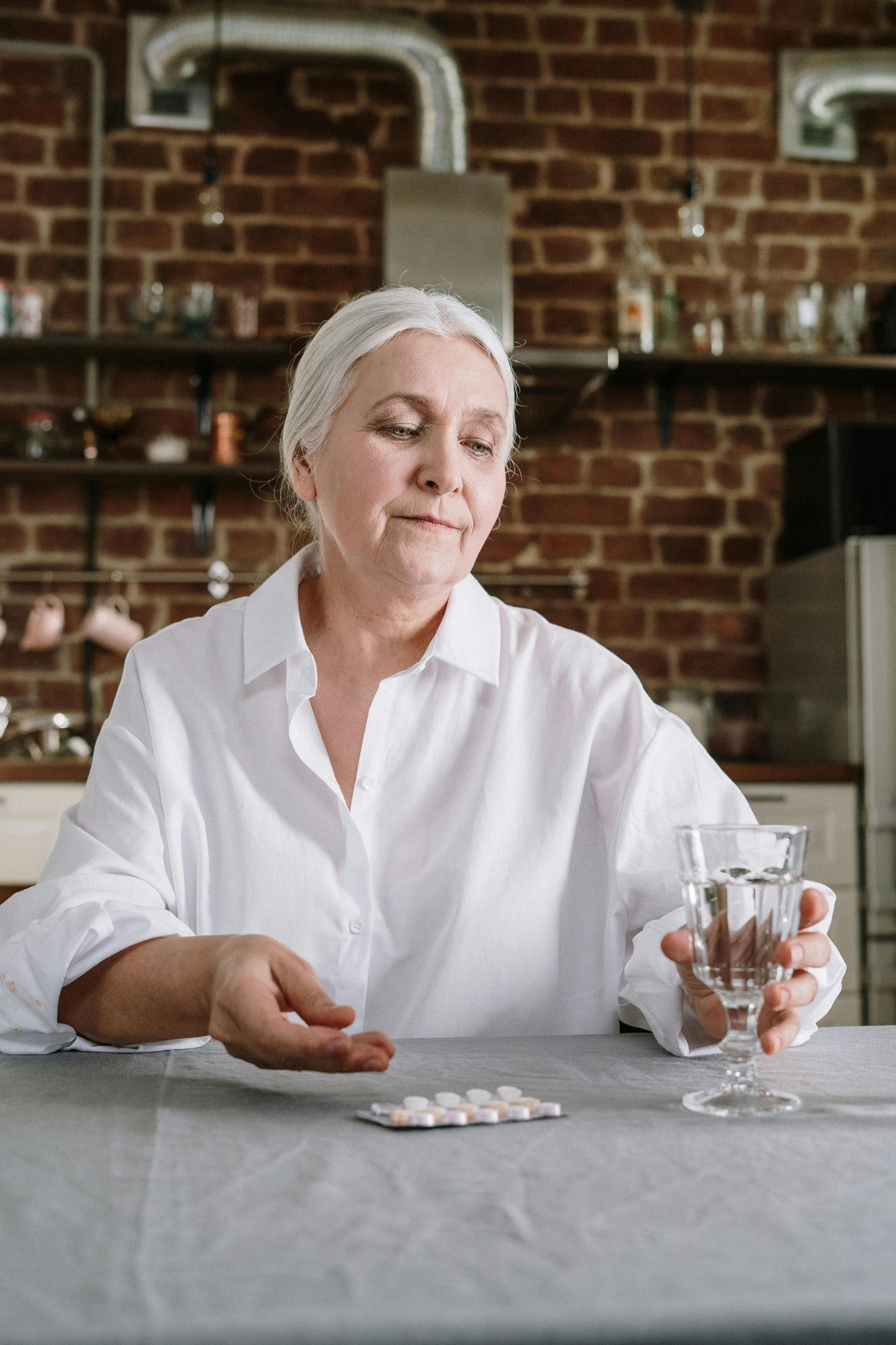 Person in a white shirt holding a glass and looking at pills on a table in a brick-walled room