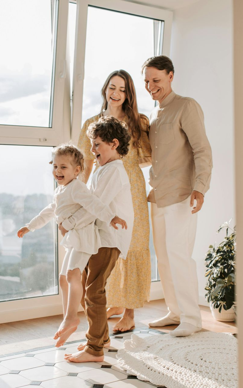 Family standing by a bright window, with two children in light clothes and two adults nearby.