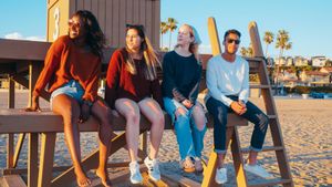 Four friends sit on a bench by the waterfront at sunset, chatting in casual clothes.