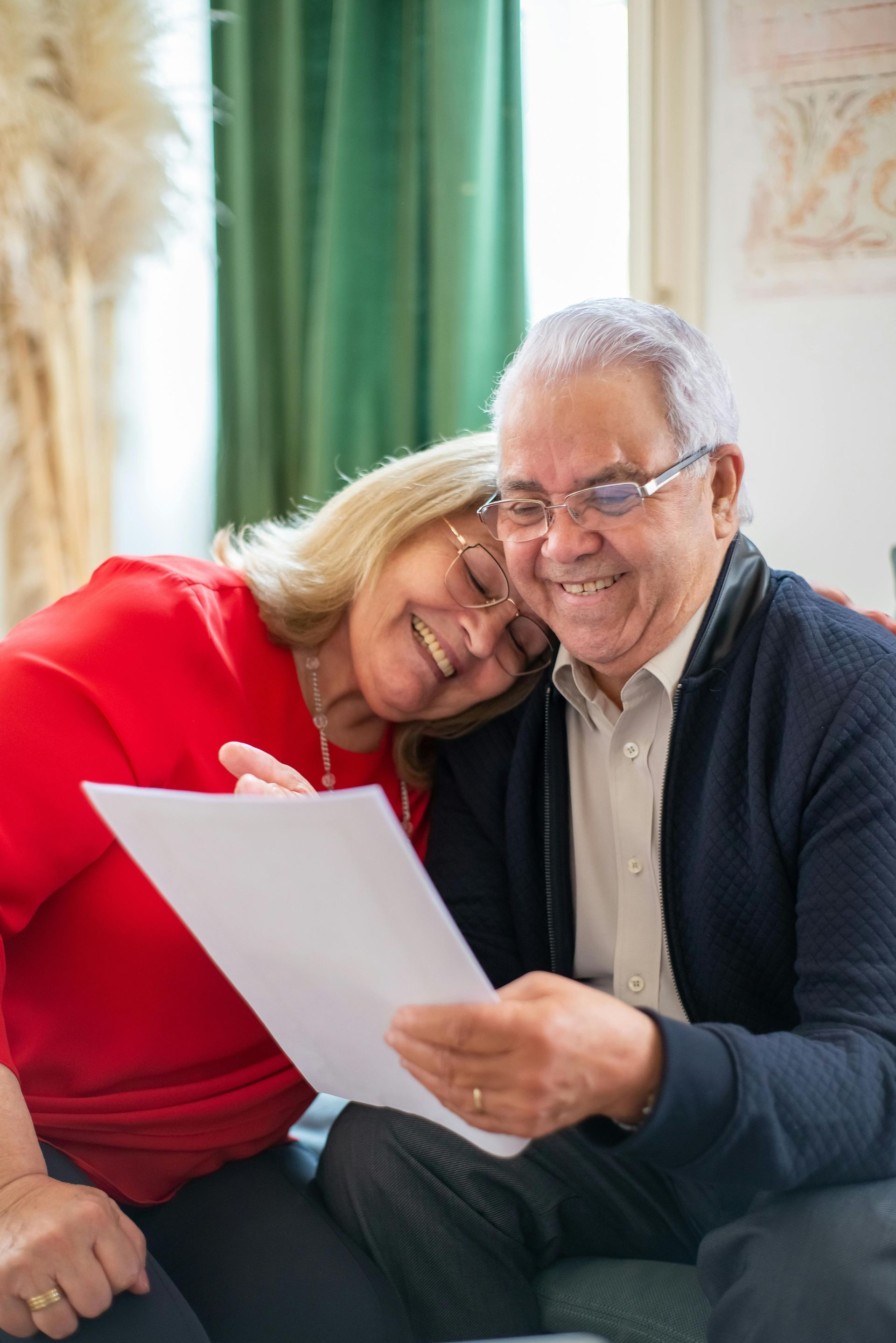 Two people smiling and reading a document together on a couch, with green curtains in the background.