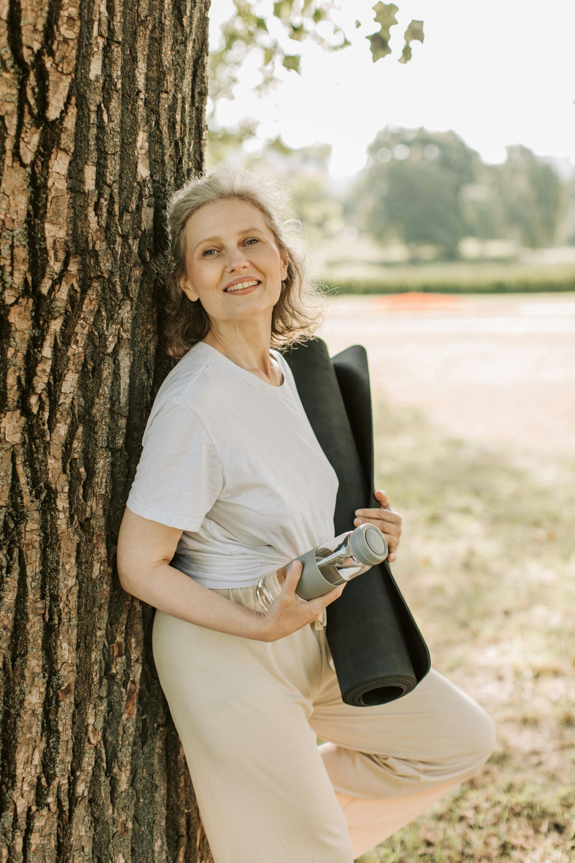 Smiling woman leans against a tree outdoors, holding a rolled yoga mat and water bottle.