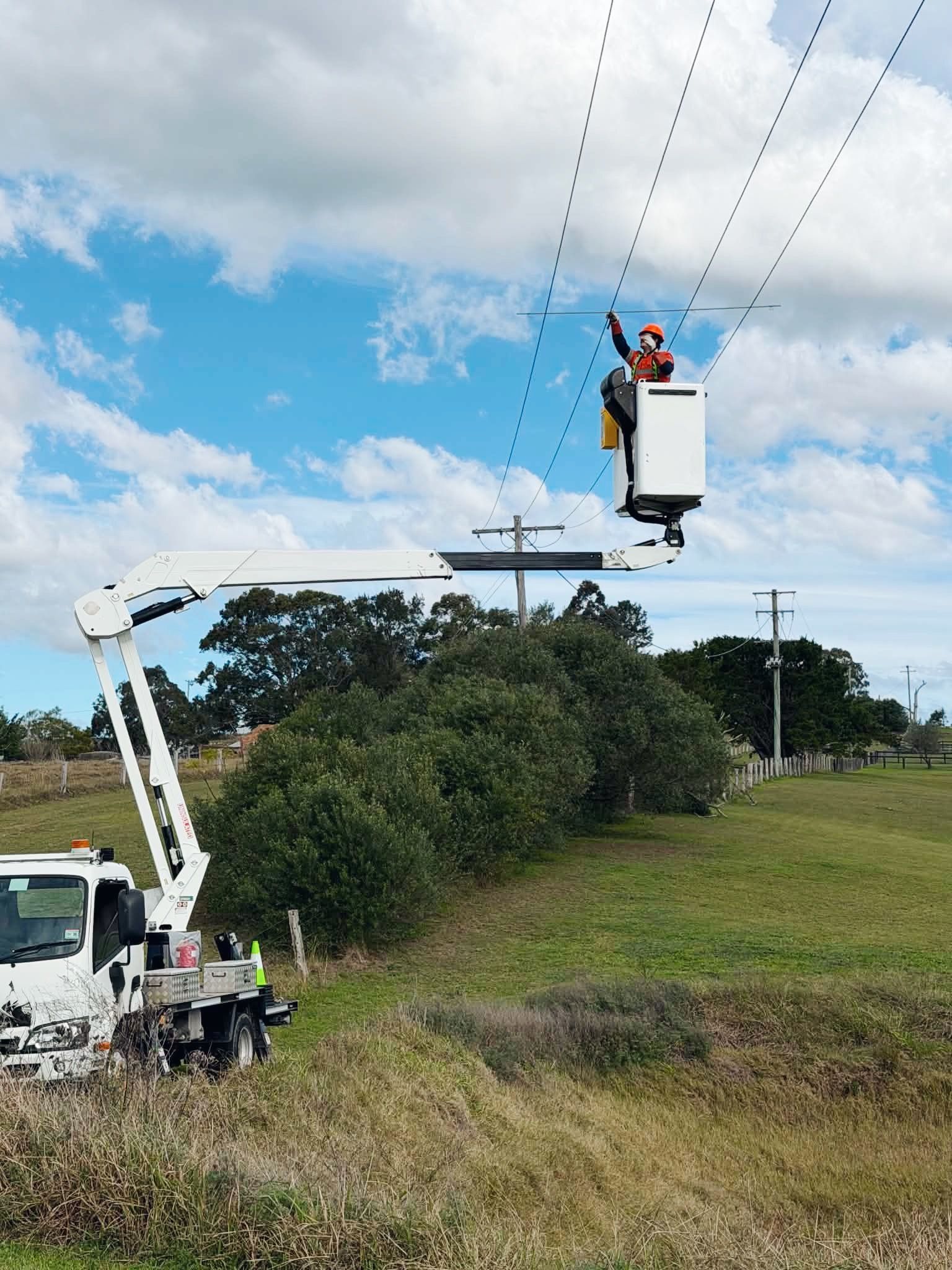 Utility worker in bucket lift inspecting power lines beside a rural roadside under a blue sky — DMP Electrical Contractors In Mandalong, NSW