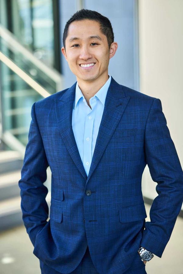 Man in blue suit, smiling with hands in pockets, standing near building.