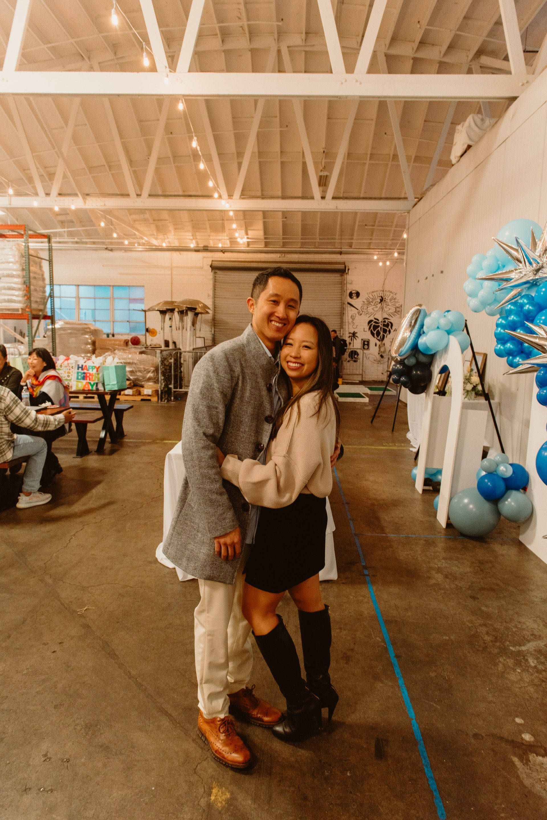 Couple embracing indoors, smiling. Man in coat, cream pants, brown shoes; woman in sweater, skirt, boots. Party decorations visible.