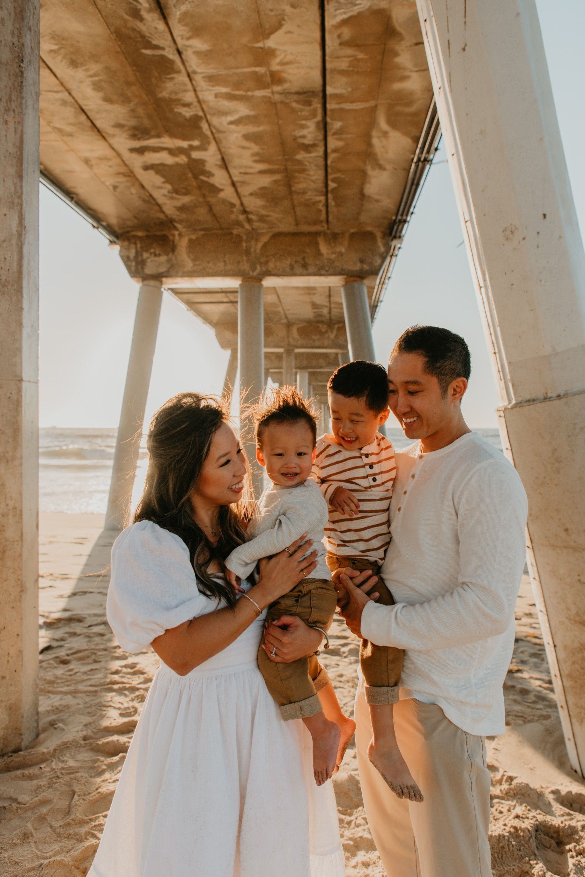 Family under pier: parents holding two young children on a sunny beach, smiling.