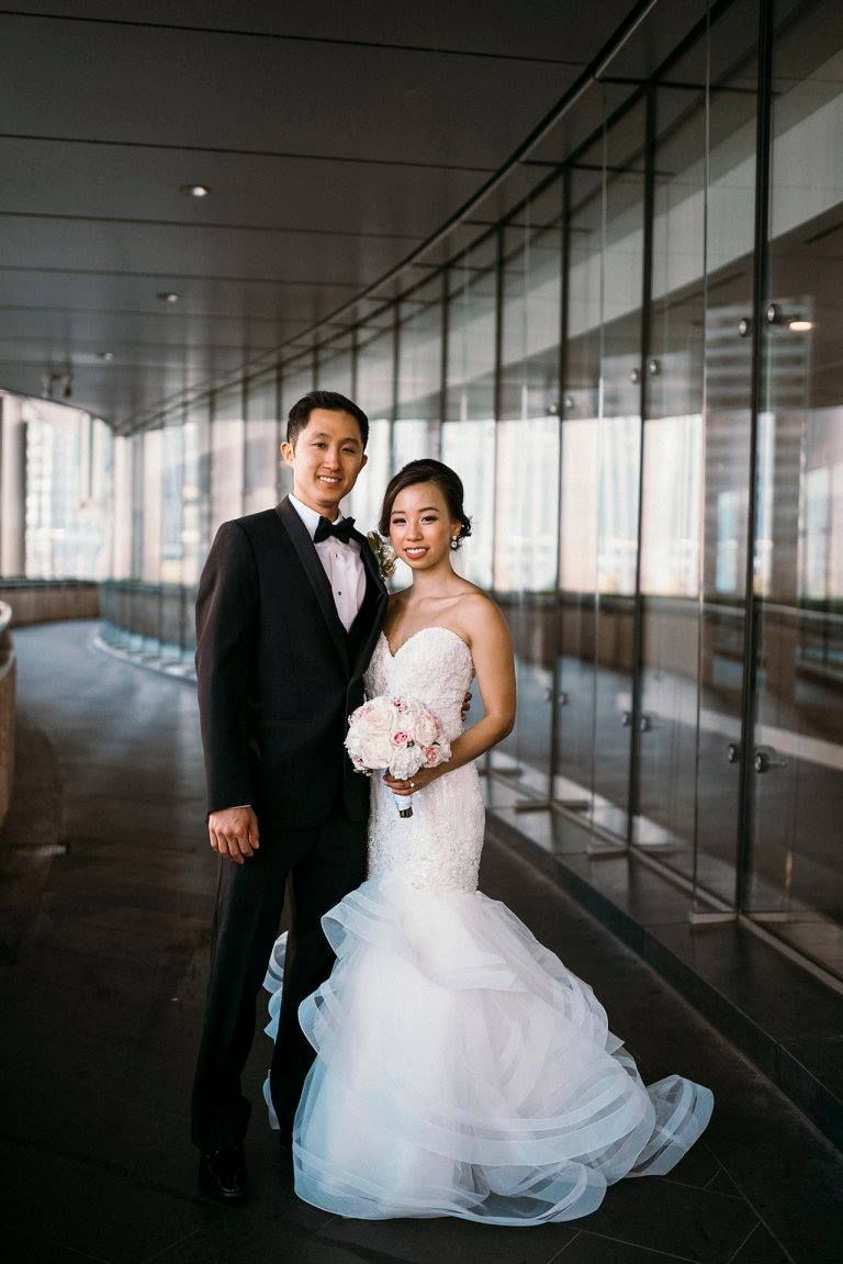 Bride and groom in formal attire posing in a modern hallway; the bride holds a bouquet.