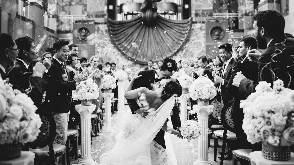 Newlyweds kiss under confetti at a wedding ceremony; guests cheer.