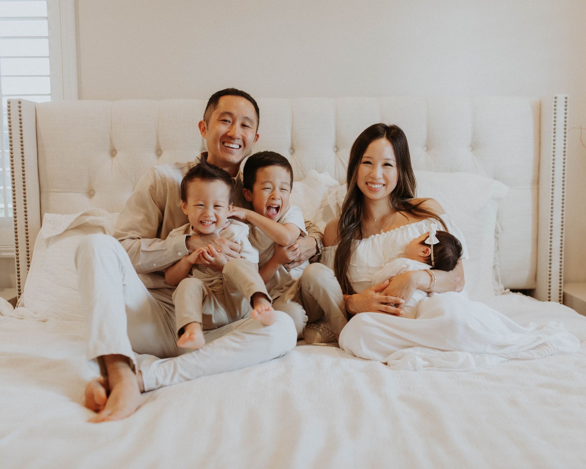 Family of five on a bed; parents, three children, one infant. Smiling faces, light-colored clothing, bright room.