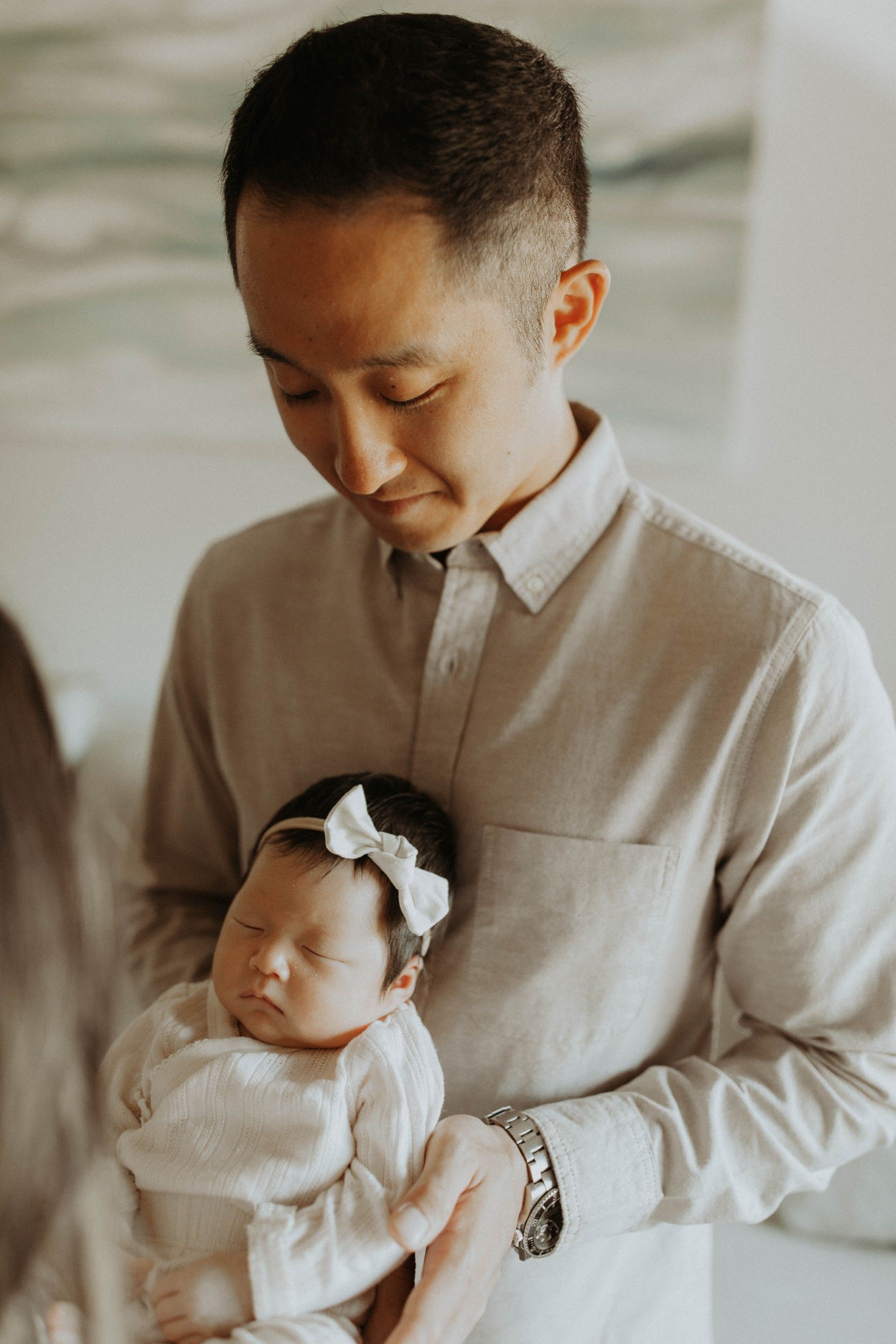 Man holding a sleeping baby wearing a white bow. They are indoors.