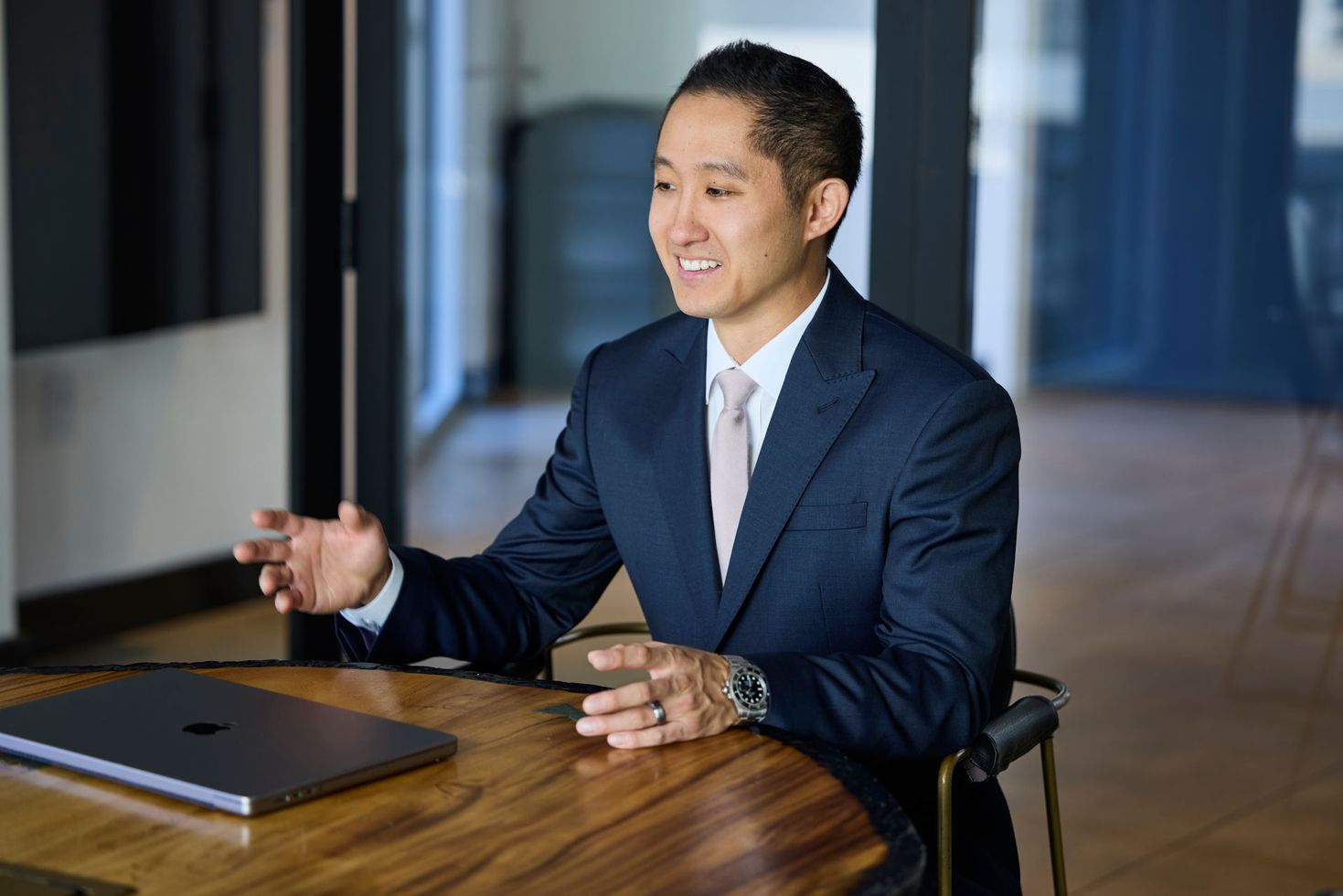 Man in a suit gestures while seated at a wooden table with a laptop in a modern office.