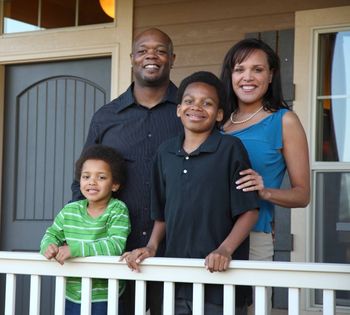 Family of four smiling on porch; house in background.