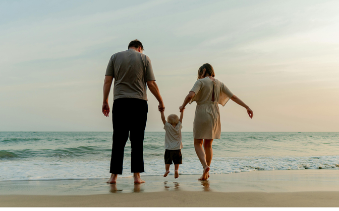 Parents holding hands with a young child, walking on a beach at sunset.