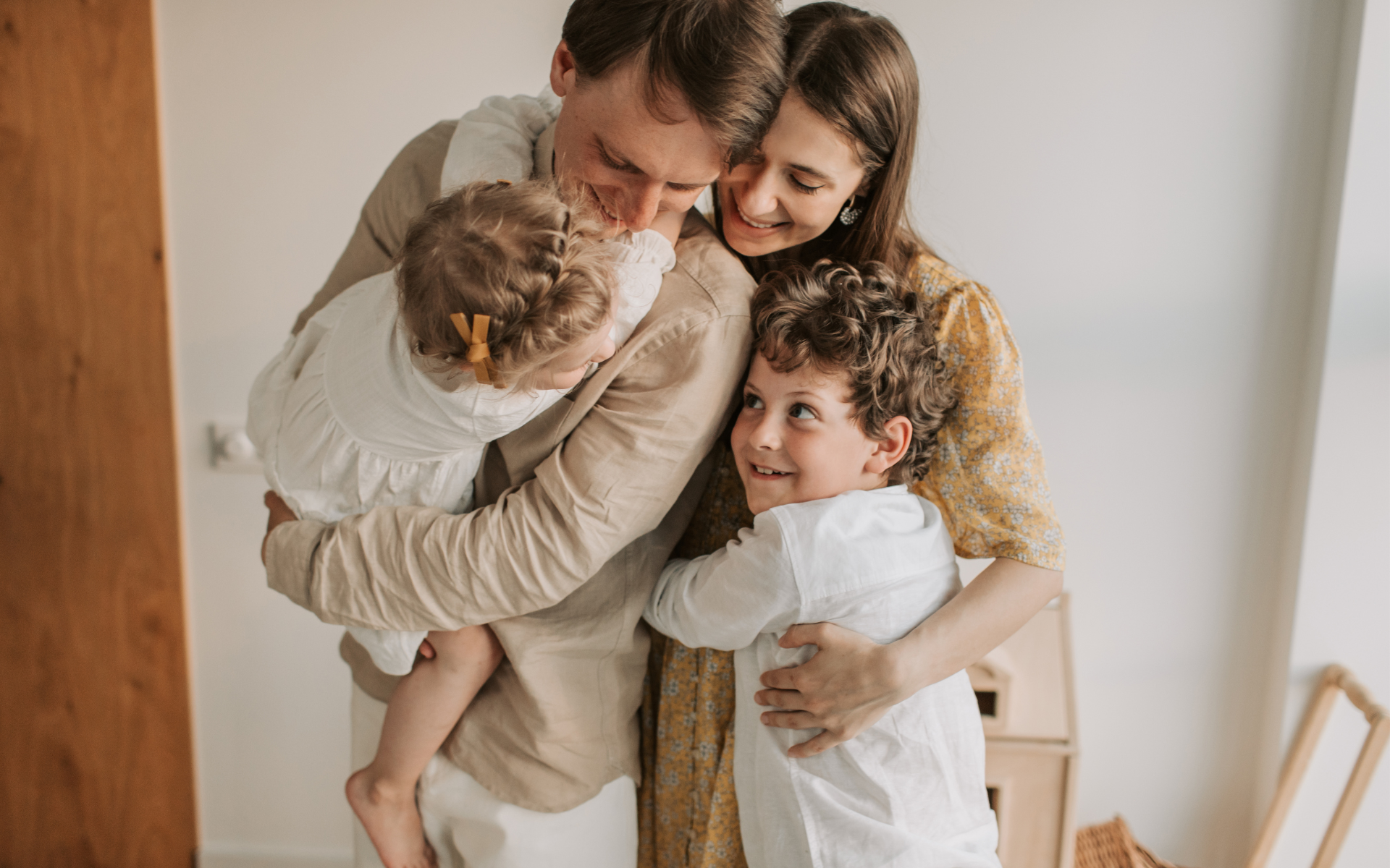 Family embracing indoors; beige and white clothing, happy expressions.
