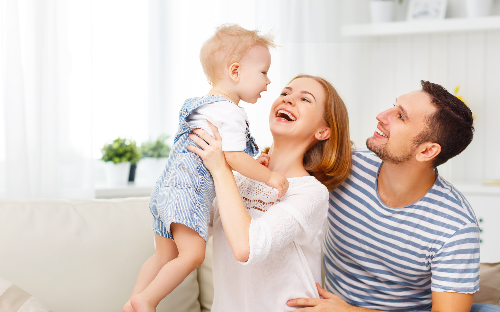 A smiling woman holds a child, a man smiles next to them, on a couch in a bright room.