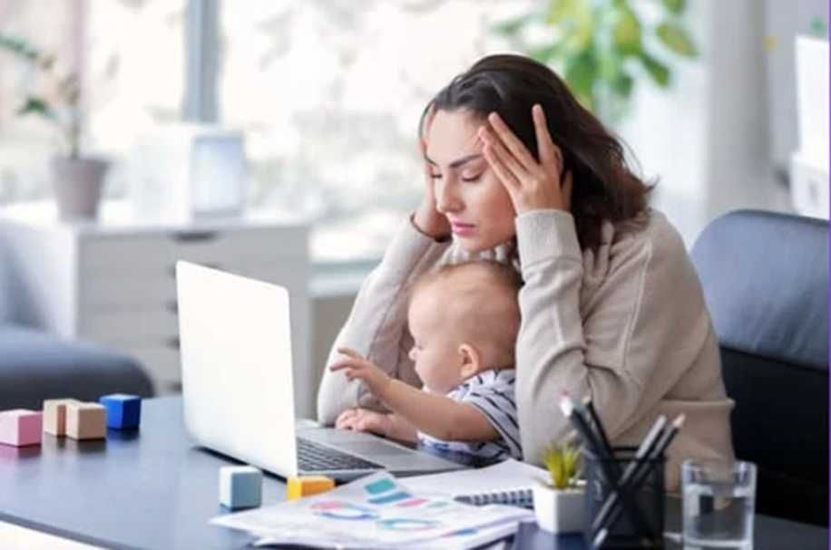 Woman with head in hands, baby on lap, working on laptop at desk.