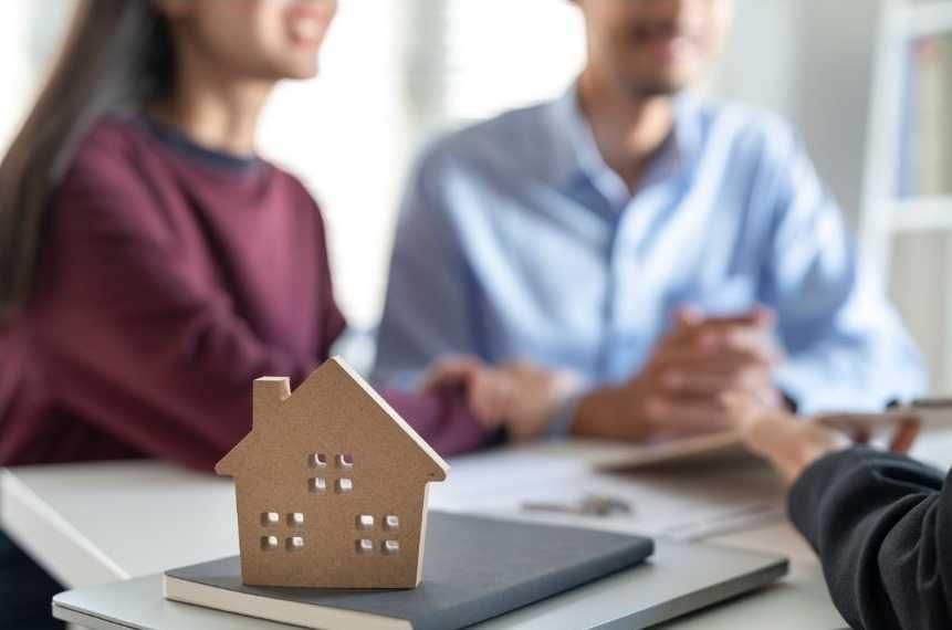 Wooden house model on a table with a couple and a person, possibly during a real estate meeting.