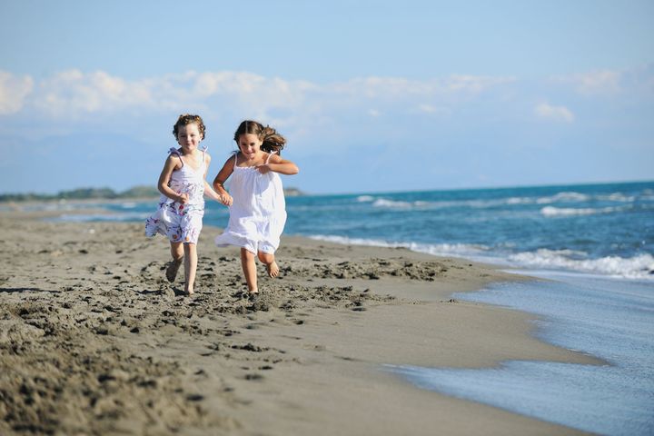 Two girls running on a sandy beach near the ocean. They wear white dresses and smile, with a blue sky and water.