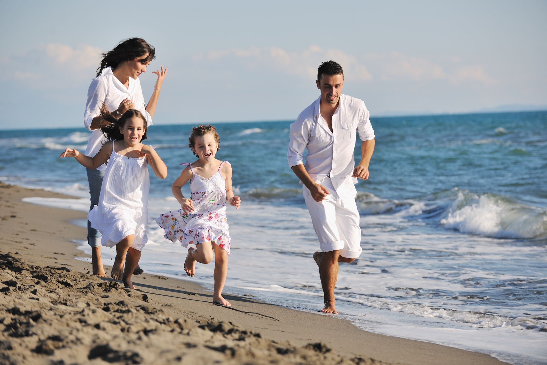 Family running on a sandy beach near the ocean; they wear white clothing and smile.