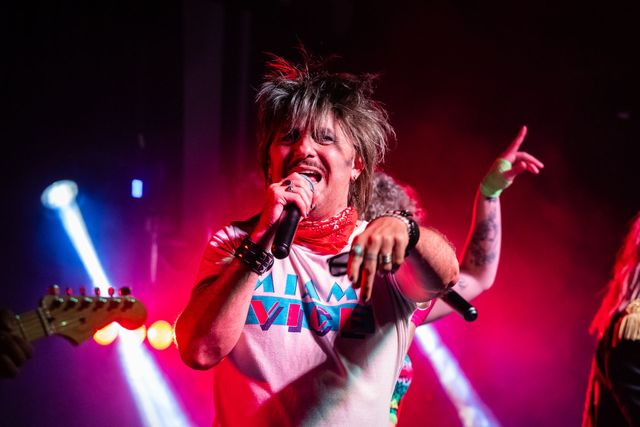 A person with spiky hair and bandana sings into a microphone on stage, gesturing with their hand — Jack West Photography In Kawungan, QLD