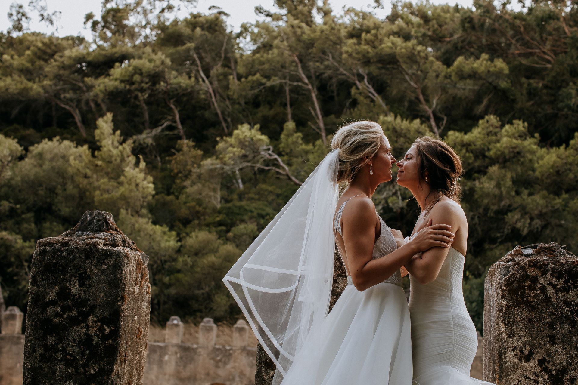 Two brides sharing a kiss outdoors, surrounded by nature, one wearing a veil.