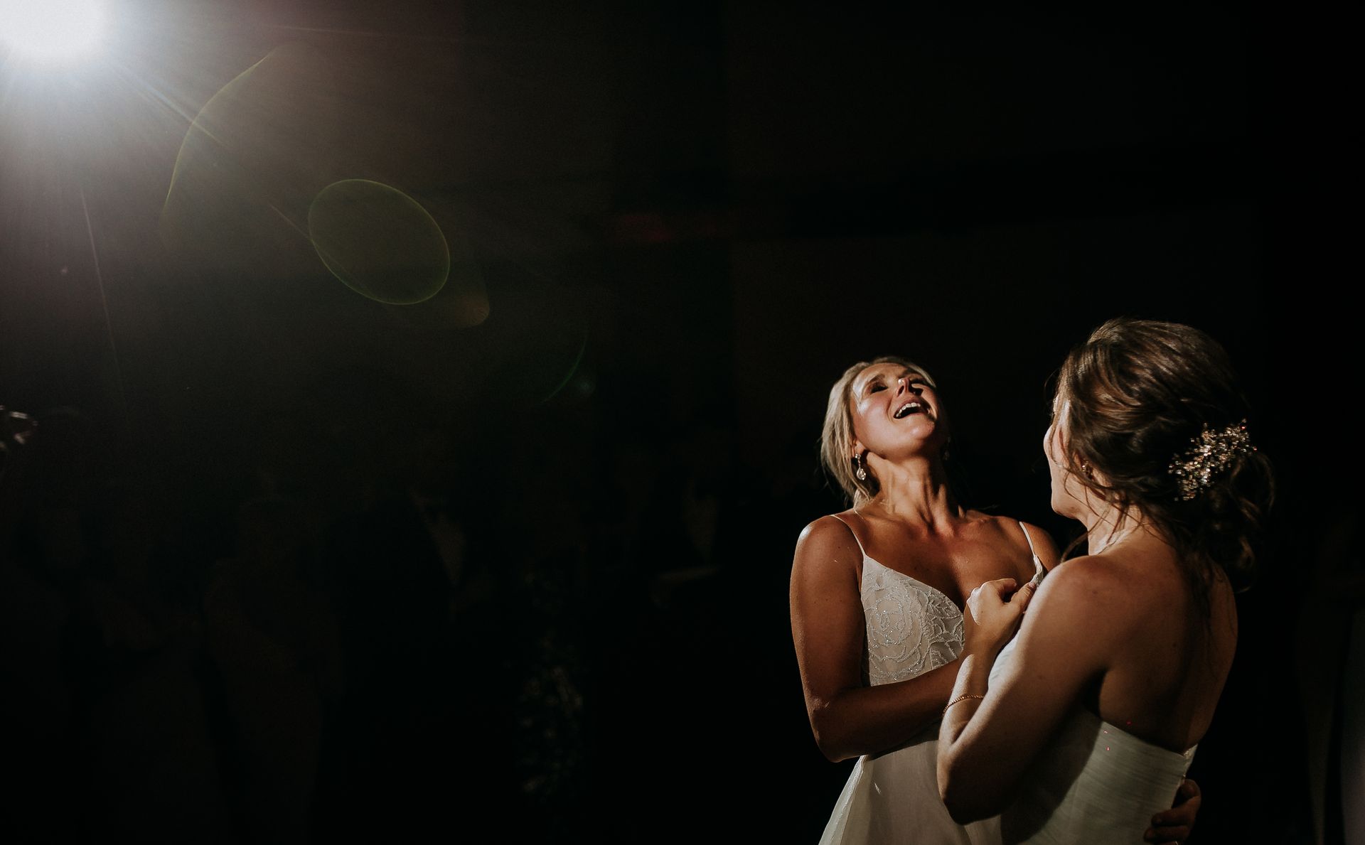 Two brides in white dresses are embracing and laughing joyfully on a dimly lit dance floor, celebrating at their wedding reception.