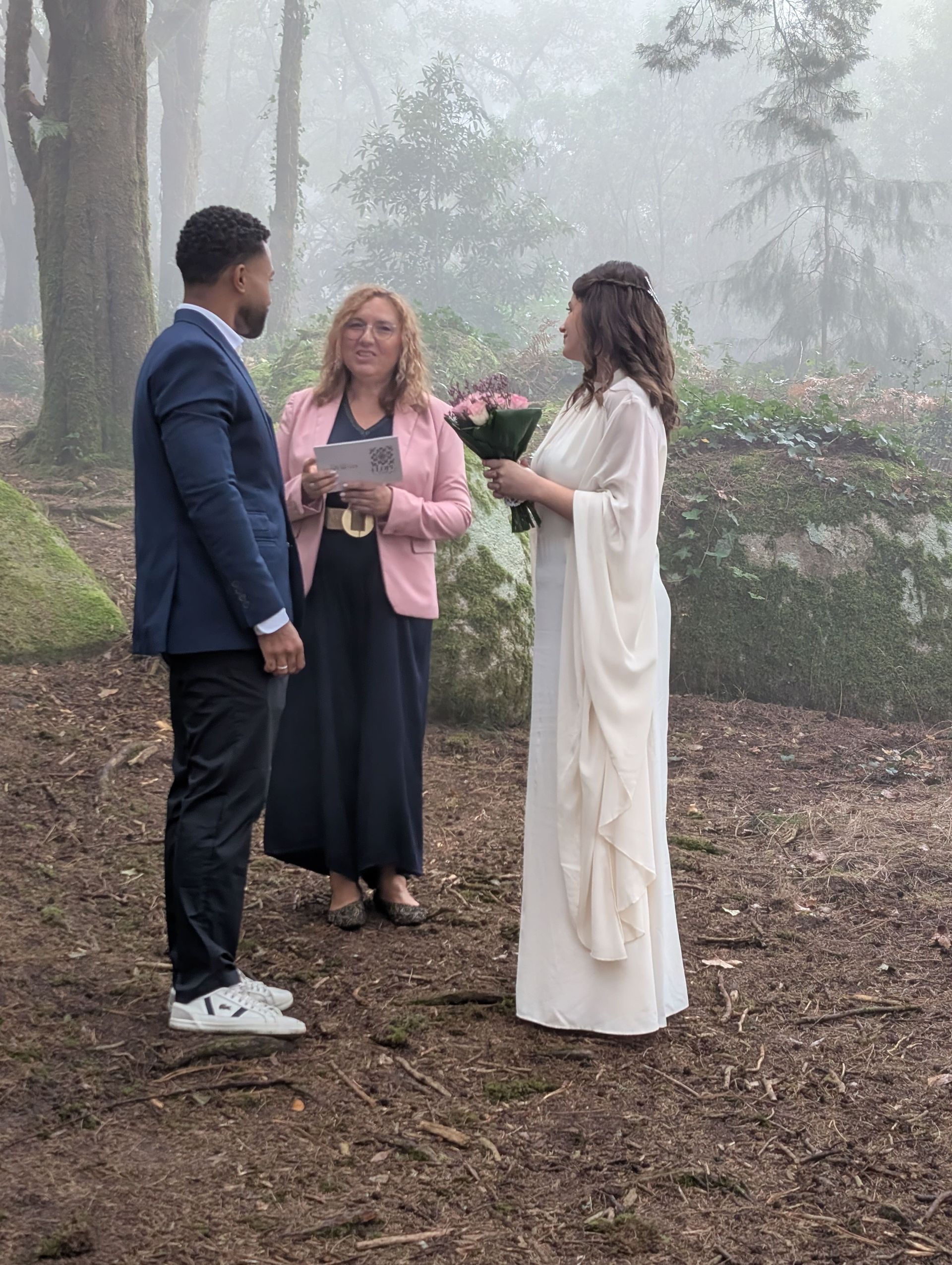Casal a celebrar um elopement na floresta da Serra de Sintra – Peninha, envoltos em neblina, com uma celebrante entre eles; a noiva segura um ramo de flores e usa um vestido fluido, criando um ambiente íntimo e místico.