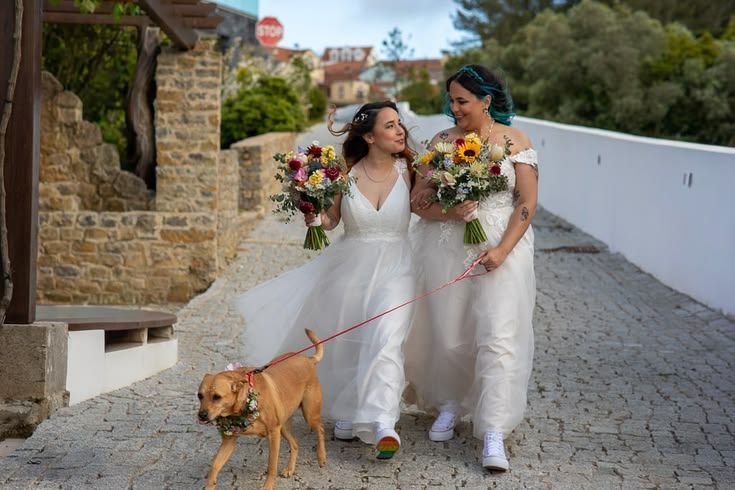 Two brides walking hand in hand along a stone path with their dog, carrying colourful bouquets.