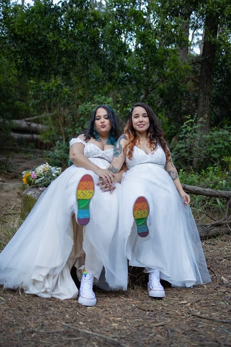 Two brides sitting together in a forest, showing off their rainbow-coloured wedding shoes.