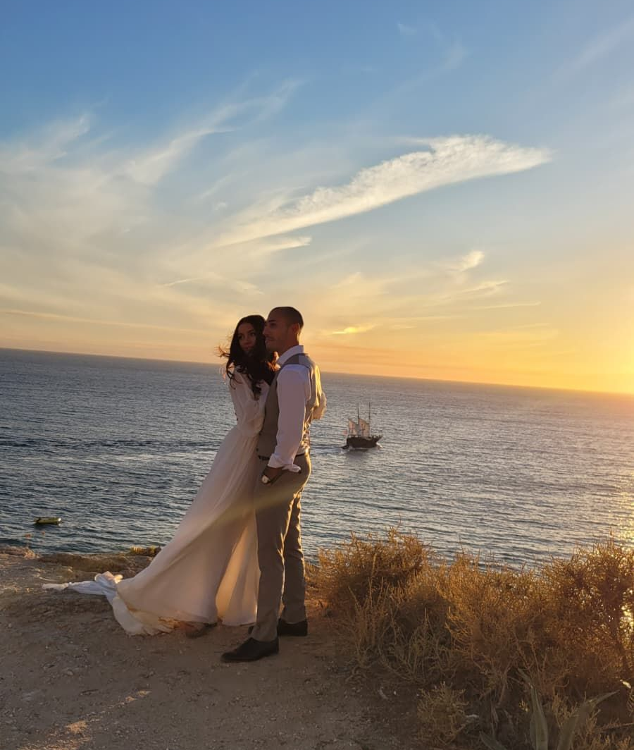 A couple embracing during their photoshoot at sunset on the coastal cliff edge in Carvoeiro, Algarve. The warm light casts a golden glow over the scene, which overlooks the ocean and a small sailboat visible on the water.