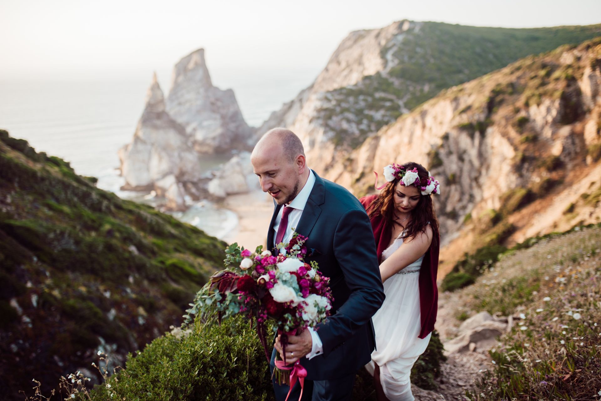 A newlywed couple is seen walking away from Praia da Ursa after their elopement ceremony. The groom, in a dark suit, is carrying a large bouquet of deep red and pink flowers. The bride wears a white dress and a floral crown. Dramatic coastal cliffs and rock formations form the backdrop.