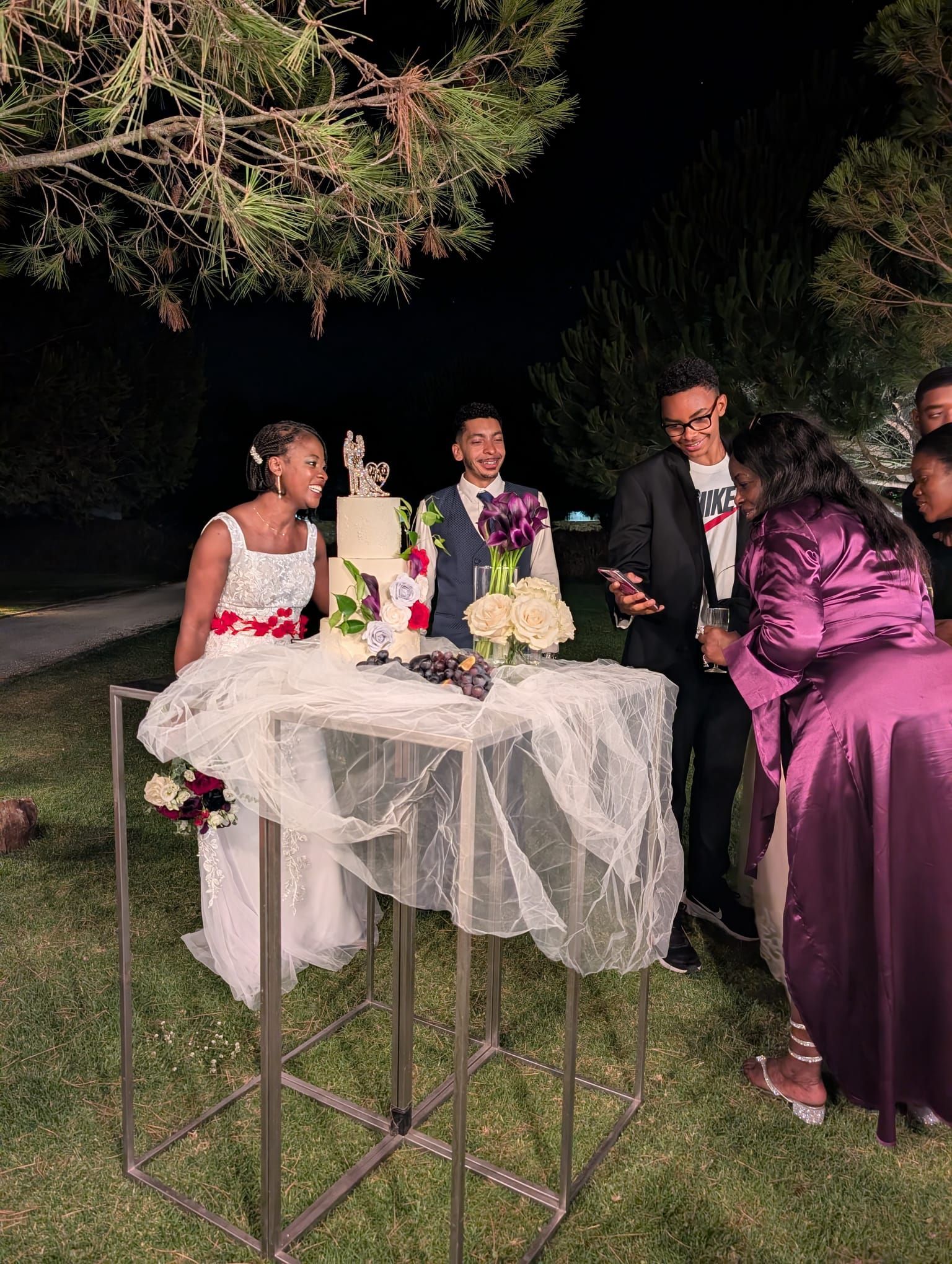 A couple and a celebrant are gathered around a table covered with sheer white fabric, illuminated by warm lights at a nighttime outdoor reception. They are preparing for the cake cutting ceremony.