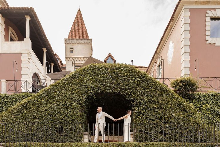 An elopement photoshoot at the Six Senses Hotel Douro Valley. A bride and groom are standing on a balcony, holding hands. The balcony railing is completely covered with sculpted green foliage that frames a pink-and-white architectural tower of the hotel behind them.
