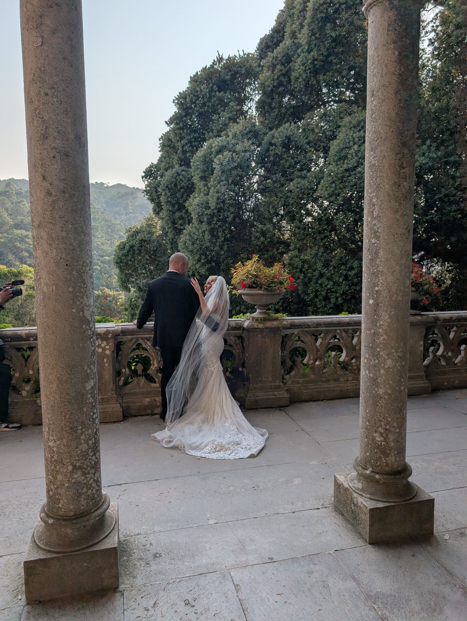 A stunning wedding photoshoot at Palácio de Monserrate in Sintra. The couple is seen from the back, standing on a classical stone balcony. The bride's long, detailed gown trails elegantly on the floor as they look out at the surrounding landscape.