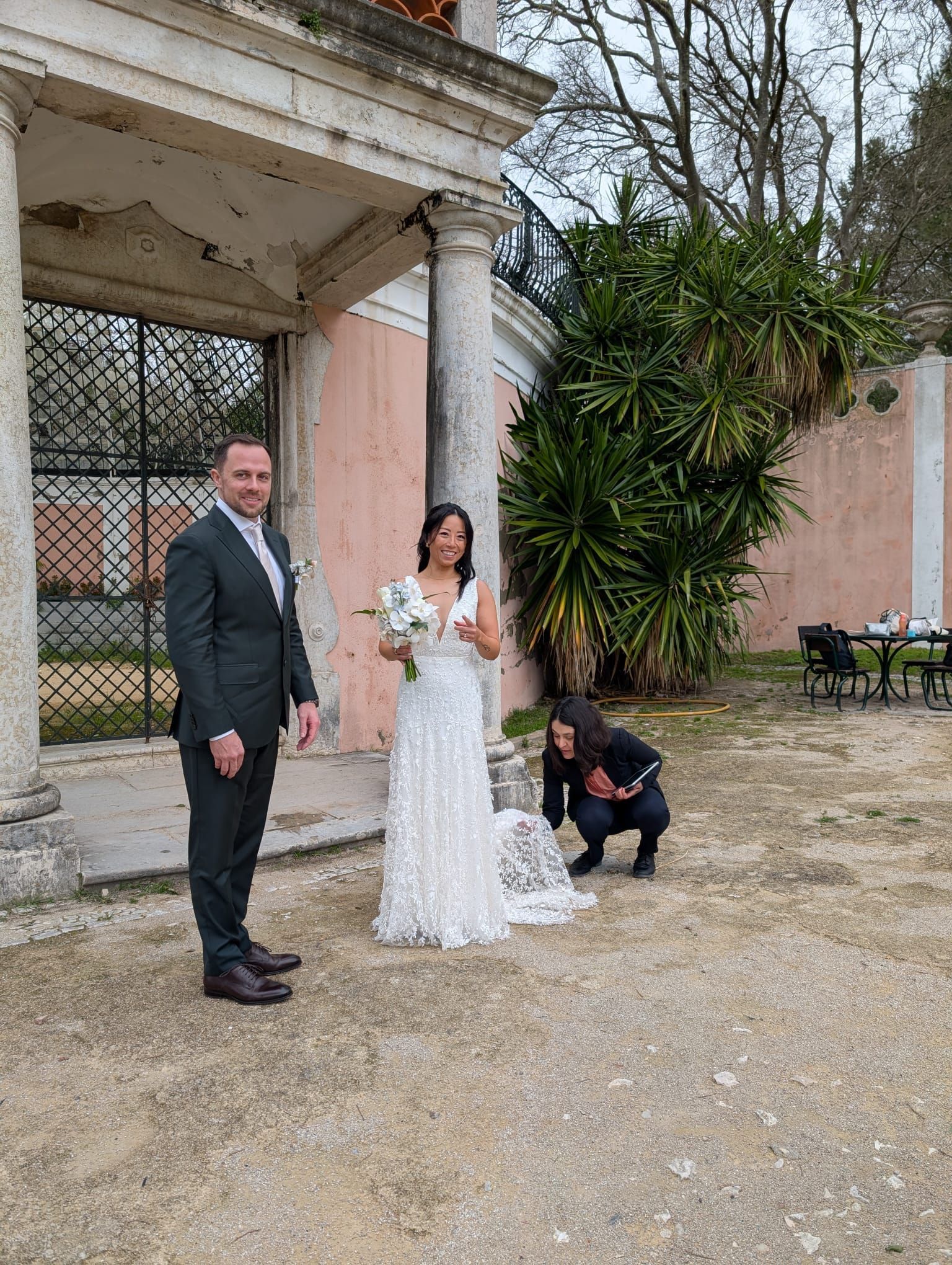 Bride and groom pose outside a historic pink building while the celebrant adjusts the bride’s dress.