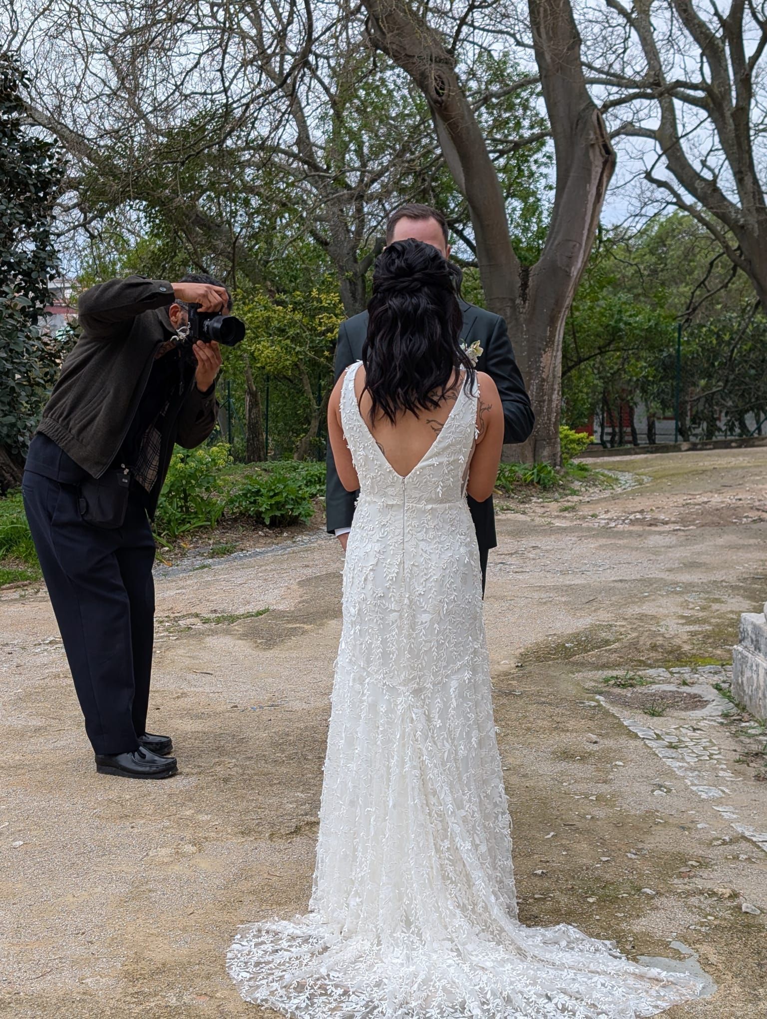 Bride in lace gown poses for photos with groom during an outdoor shoot in a wooded garden.