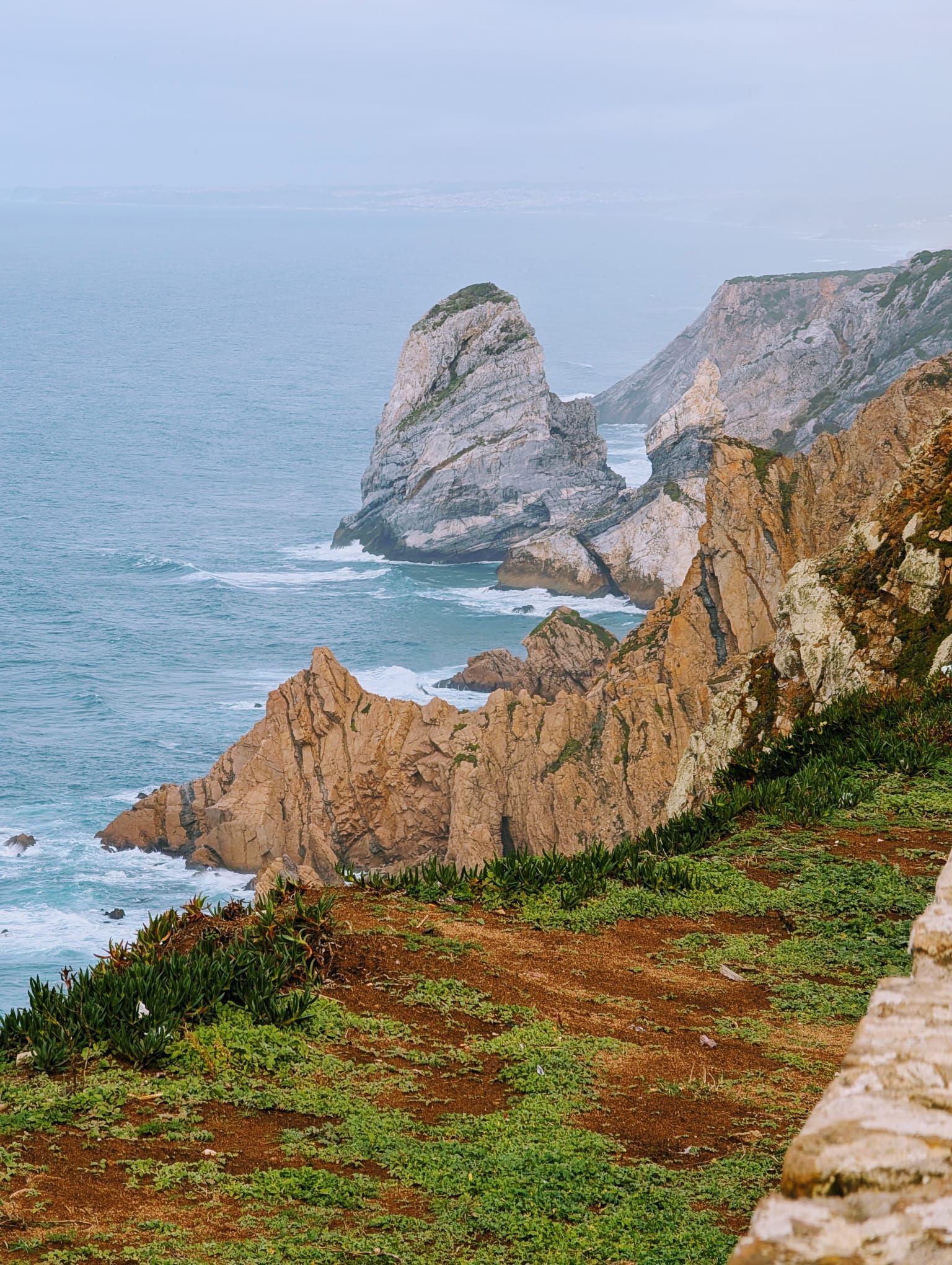 A dramatic view of rugged coastal cliffs dropping into the deep blue ocean, with green and reddish scrub on the clifftops. Location Cabo da Roca.