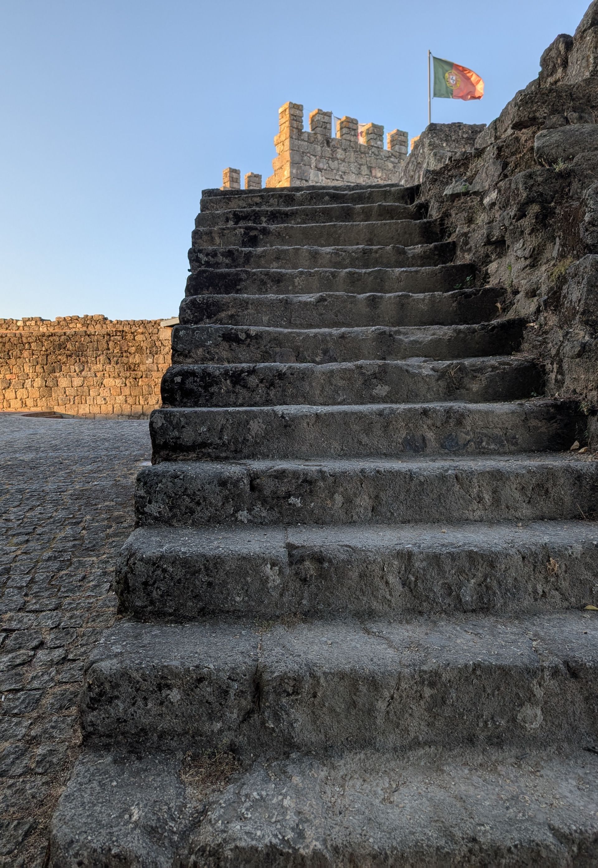 A dramatic view up a set of rough stone stairs leading along the high wall of the Castelo Branco castle. A Portuguese flag flies prominently from the top of the fortress wall. The text