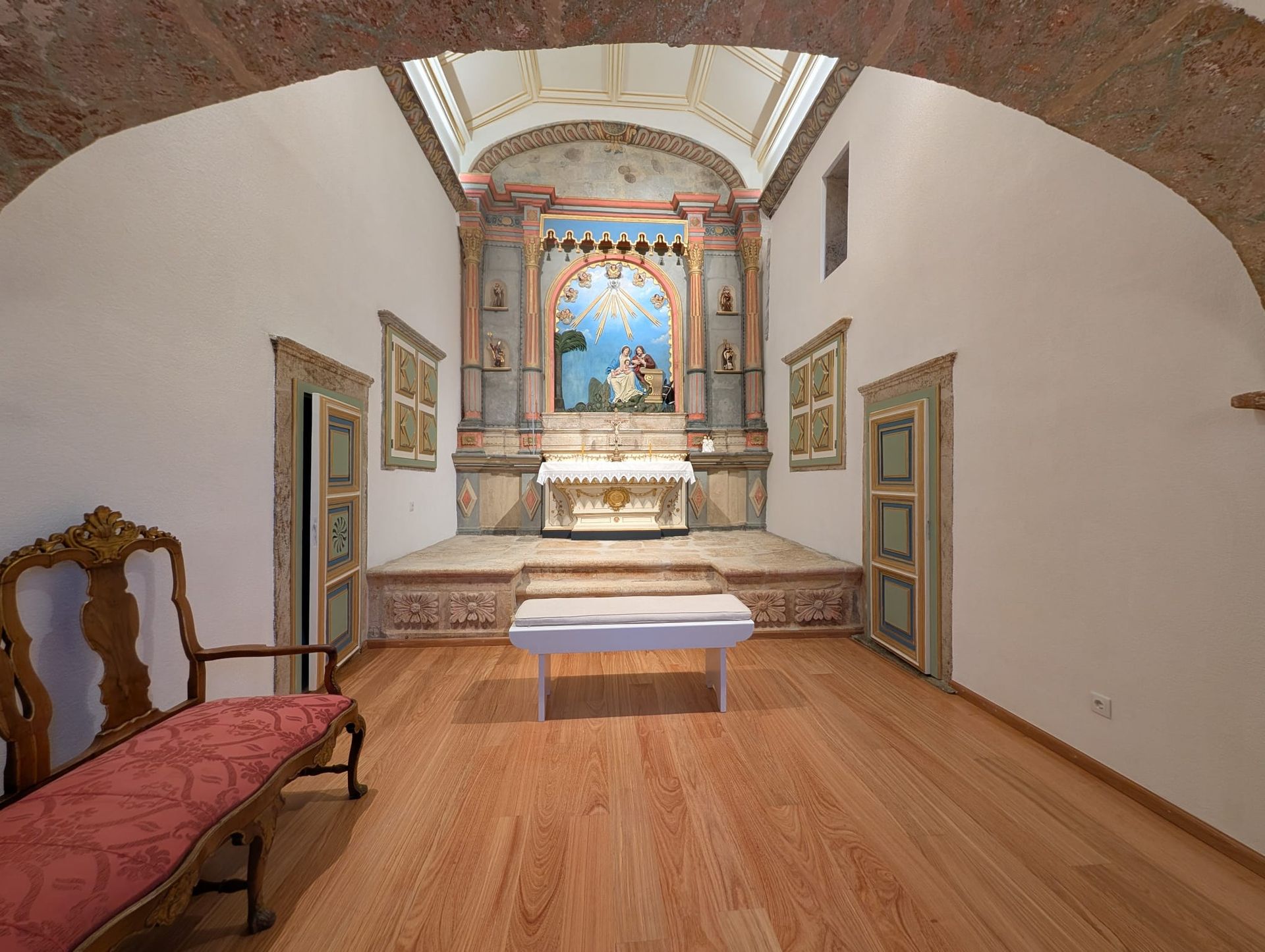 The interior of the small, beautiful Chapel of Hotel Quinta do Barrilário, where Catholic wedding ceremonies can be held. The space is ornate, featuring an altar with a religious image, bright decorative walls, and polished wooden floors.