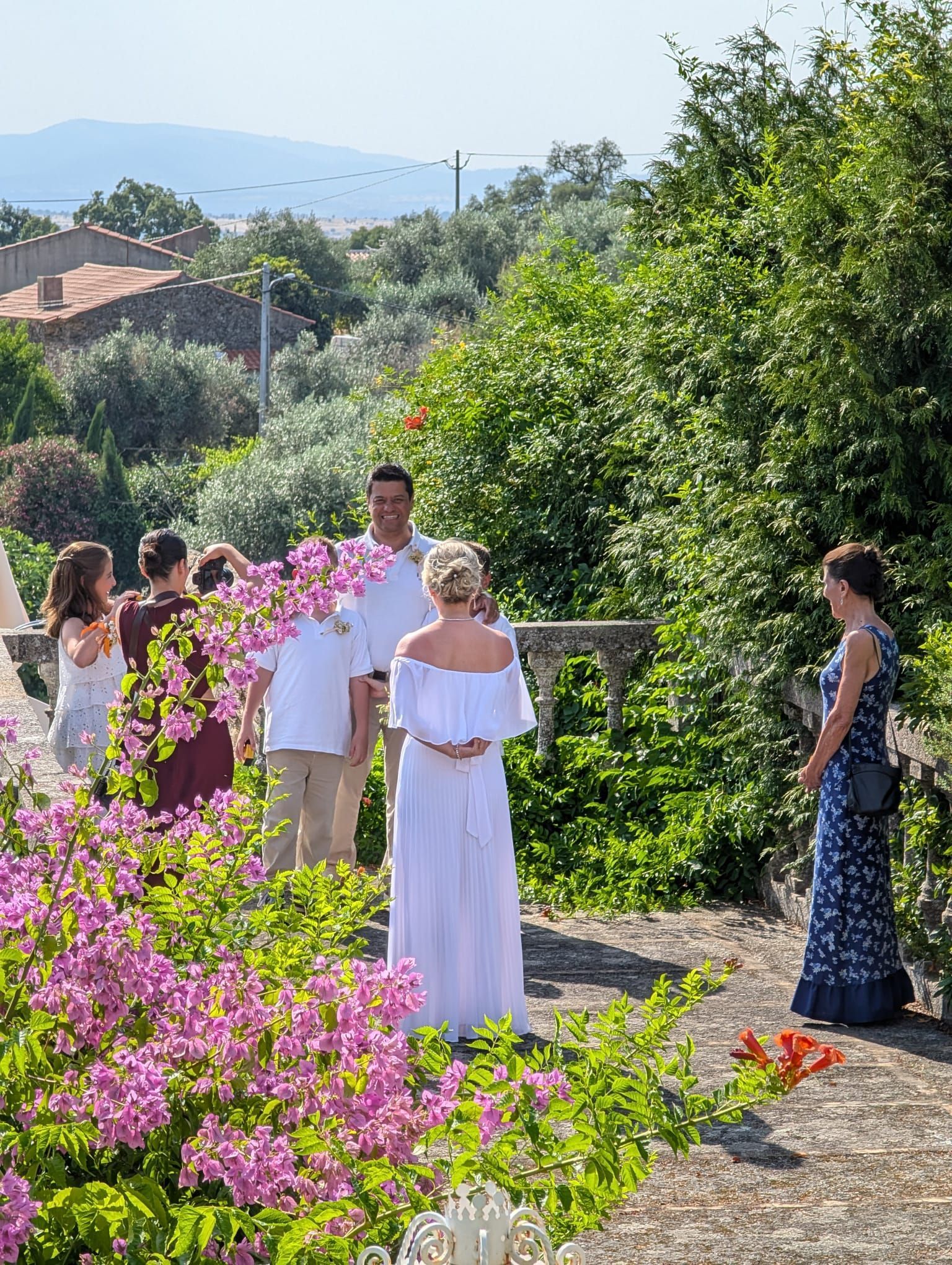 A first look at a vow renewal ceremony taking place on an XVIII century balcony in Medelim, Idanha-a-Nova. The groom and his three children (twin sons and a daughter) stand looking toward the bride. The bride's mother-in-law is visible behind the bride. The setting is beautiful, surrounded by green foliage and bright pink bougainvillea.
