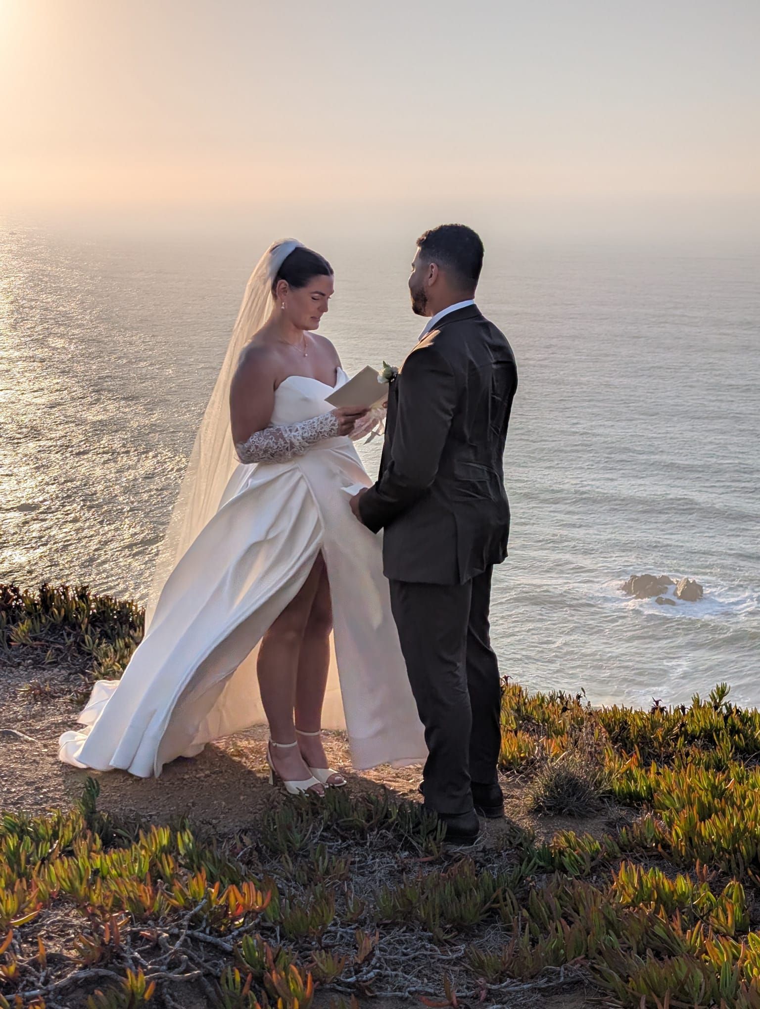 A romantic close-up of a couple at Cabo da Roca, the westernmost point of continental Europe, at sunset. The bride, in a long white gown, and the groom, in a dark suit, are holding hands and gazing at each other on a cliff edge overlooking the ocean.