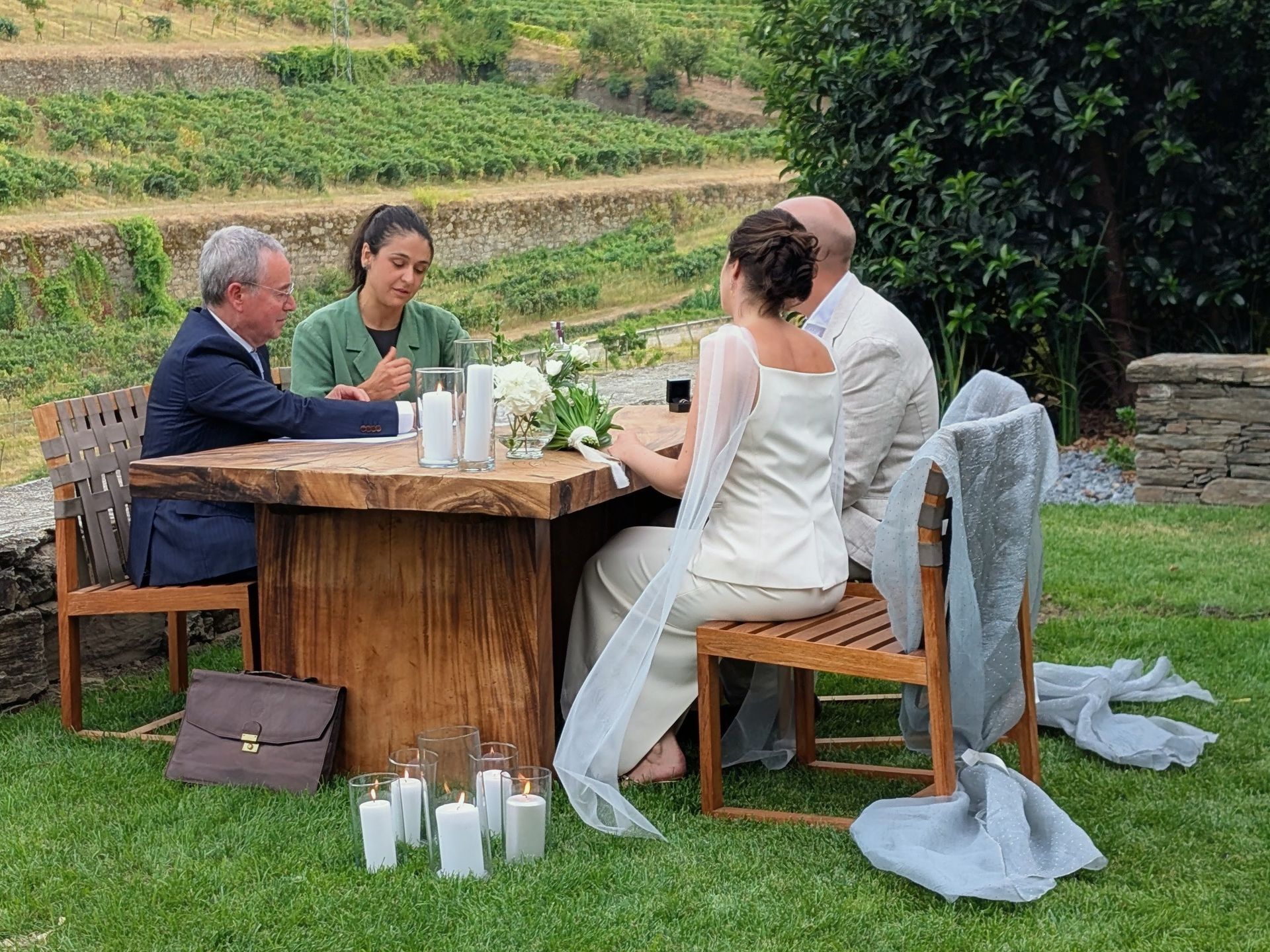 An official legal marriage ceremony is taking place outdoors at the Six Senses Hotel Douro Valley. The couple, dressed in wedding attire, sits at a rustic wooden table with the Conservador from the registry and an official translator. White candles are placed on the ground next to the table.