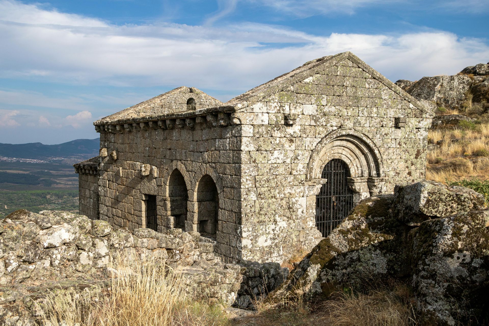 The stone ruins of a Templar chapel in Monsanto, known for its historical setting and as a filming location for the series House of the Dragon. The ancient building features rounded arches and is set dramatically on a rocky outcrop under a clear blue sky.